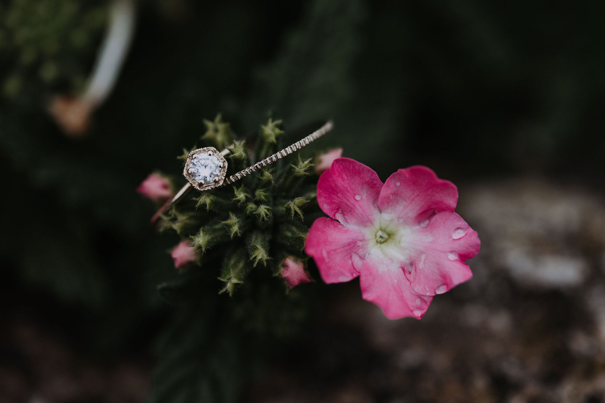 A close up photo of delicate wedding band and engagement ring on a pink flower